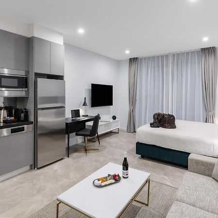 Meriton Suites Sussex Street, Сидни Екстериор снимка The photo shows a modern studio apartment. On one side, there is a kitchenette with sleek gray cabinets, a refrigerator, and built-in appliances. In the living area, there is a comfortable gray sofa, a white coffee table with a fruit platter and a bo
