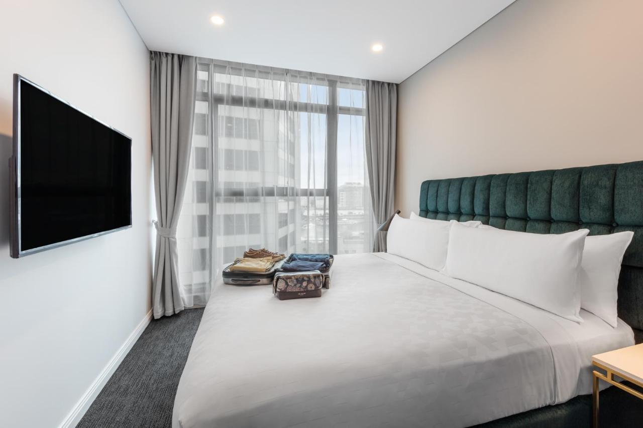 Meriton Suites Sussex Street, Сидни Екстериор снимка The photo shows a modern hotel bedroom. The room features a large bed with a green upholstered headboard and crisp white linens. There are two pillows on the bed and a few folded items of clothing neatly placed on it. A flat-screen TV is mounted on t