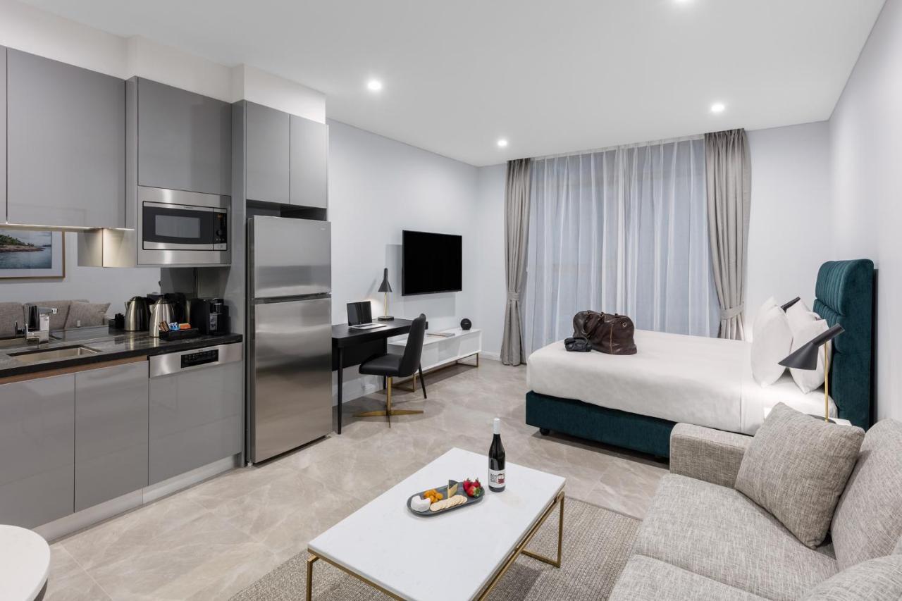 Meriton Suites Sussex Street, Сидни Екстериор снимка The photo shows a modern studio apartment. On one side, there is a kitchenette with sleek gray cabinets, a refrigerator, and built-in appliances. In the living area, there is a comfortable gray sofa, a white coffee table with a fruit platter and a bo
