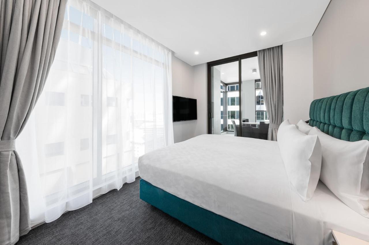 Meriton Suites Sussex Street, Сидни Екстериор снимка The photo shows a modern bedroom featuring a large bed with a white duvet and pillows. The headboard is turquoise, adding a pop of color to the neutral-toned room. There are sheer white curtains covering large windows that let in natural light, creat