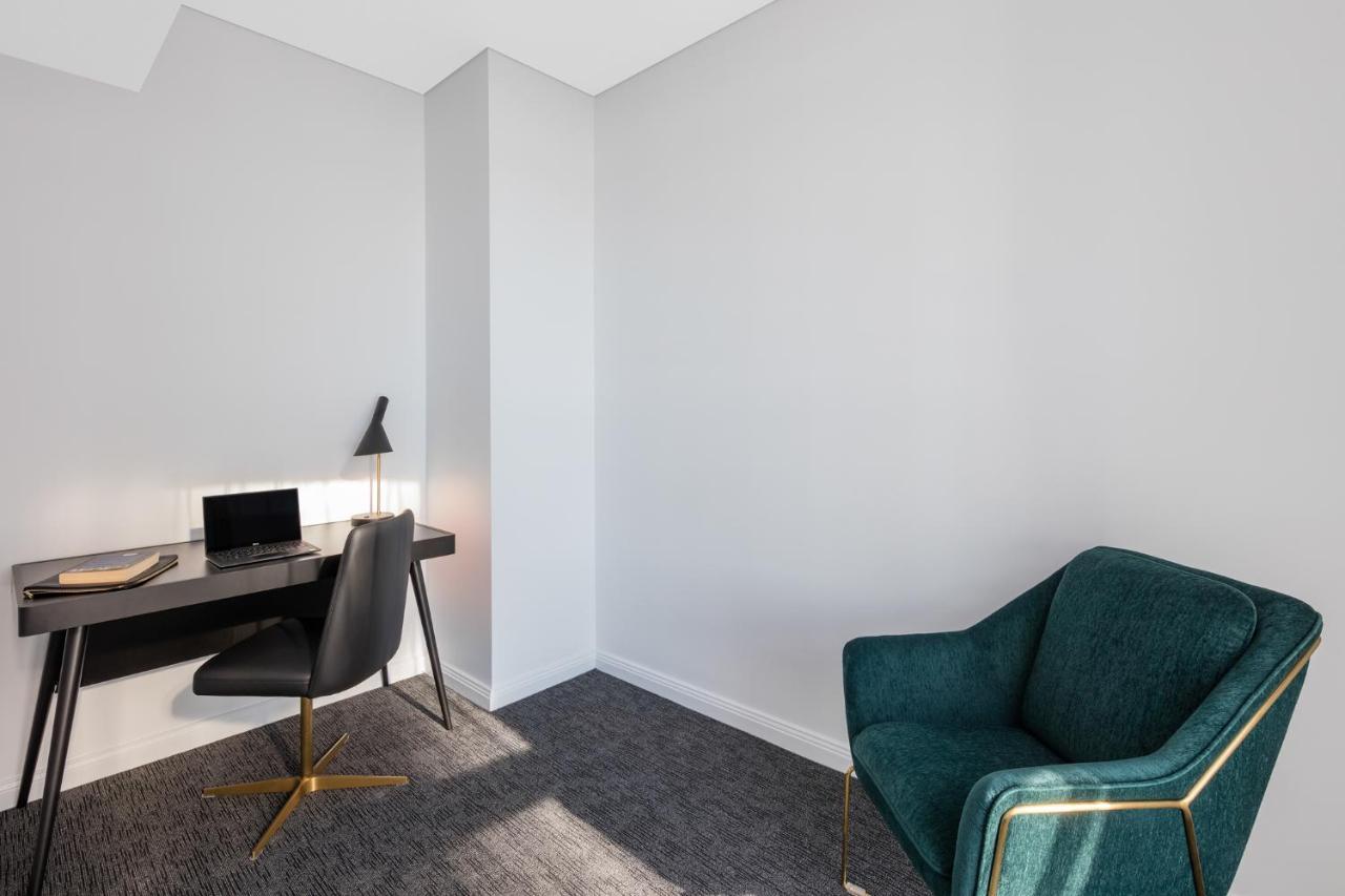 Meriton Suites Sussex Street, Сидни Екстериор снимка The photo depicts a minimalist interior space, likely a small office or study area. It features a simple, modern desk with a black lamp and a laptop or tablet on it. A sleek black chair accompanies the desk, while on the opposite side of the room, th