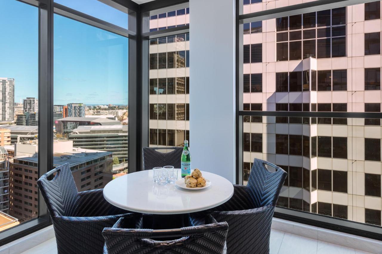 Meriton Suites Sussex Street, Сидни Екстериор снимка The photo shows a modern dining space with a round white table set for four. There are four black woven chairs around the table. On the table, there's a bottle of sparkling water, two glasses, and a small plate with several cookies. The large windows