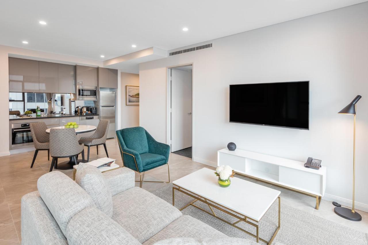 Meriton Suites Sussex Street, Сидни Екстериор снимка The photo shows a modern living room area. It features a light gray sofa and a coffee table with a decorative plant. There are two chairs, one in teal and a round one, positioned around the coffee table. A flat-screen TV is mounted on the wall, and t