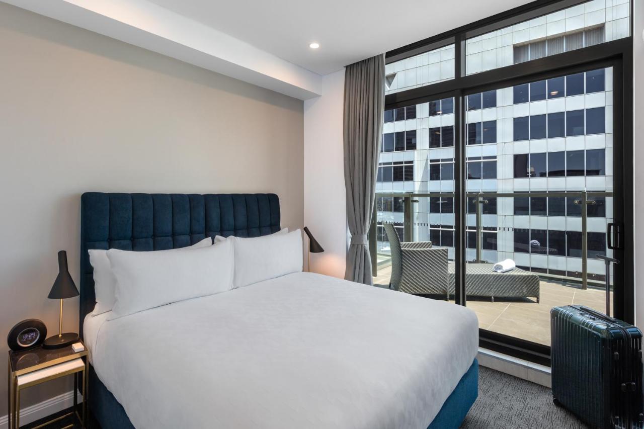Meriton Suites Sussex Street, Сидни Екстериор снимка The photo shows a modern hotel room with a large bed featuring a blue upholstered headboard and white linens. Natural light enters through a large window, revealing a view of a building exterior, likely part of the hotel or surrounding area. The room