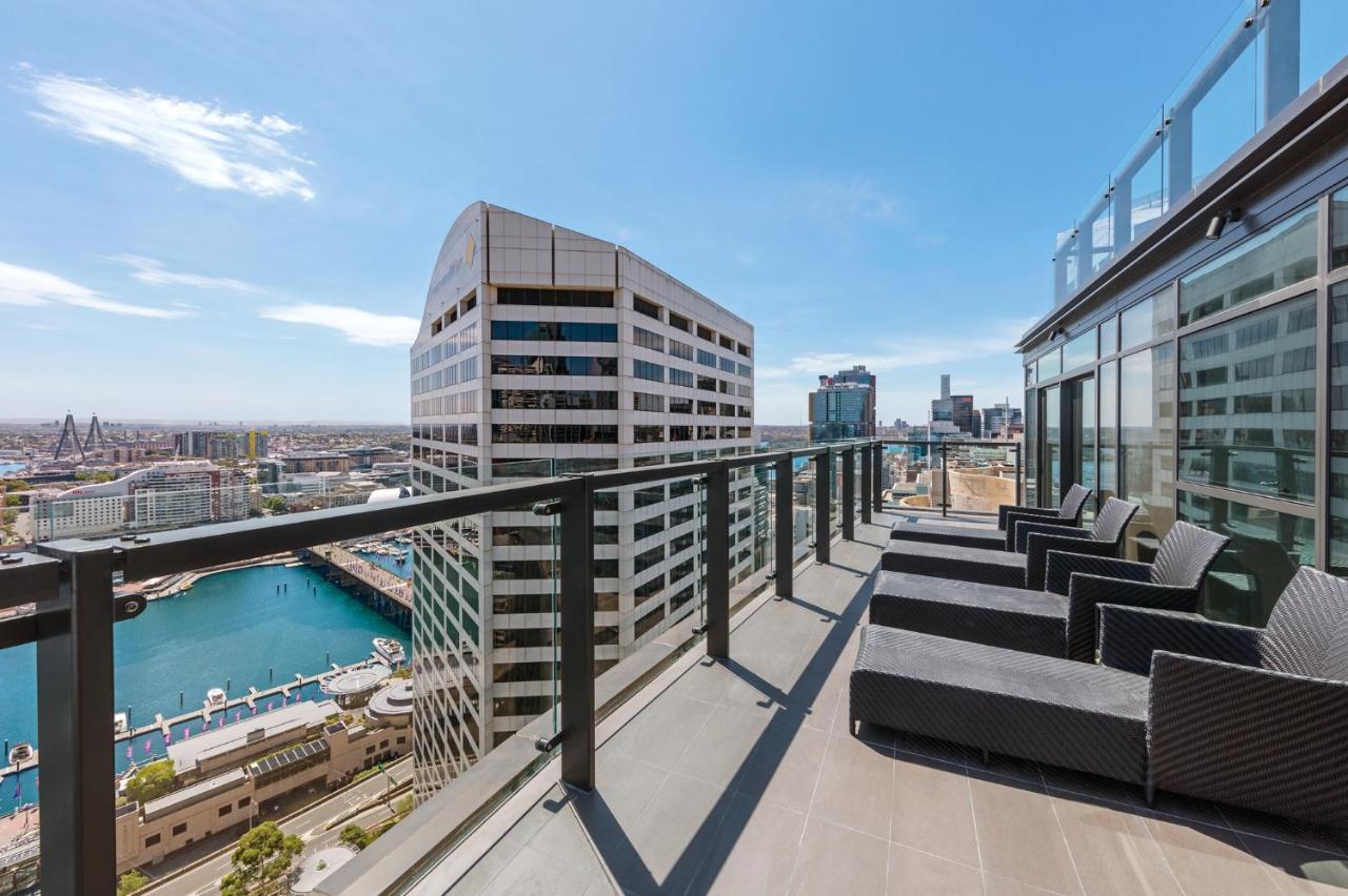 Meriton Suites Sussex Street, Сидни Екстериор снимка The photo shows a balcony or terrace with a panoramic view of a city skyline. The balcony features modern outdoor furniture, including sleek lounge chairs. In the background, you can see a waterfront area with buildings and a harbor, along with a cle