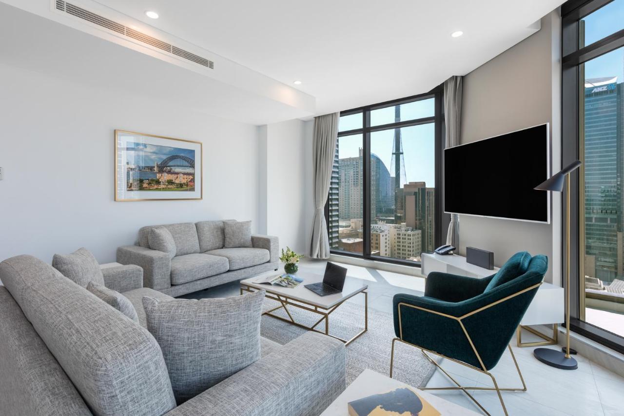 Meriton Suites Sussex Street, Сидни Екстериор снимка The photo shows a modern living room with large windows offering a view of a city skyline. The room features two gray sofas and a stylish green accent chair. A sleek coffee table is placed in front of the sofas, alongside a laptop. There’s a flat-scr
