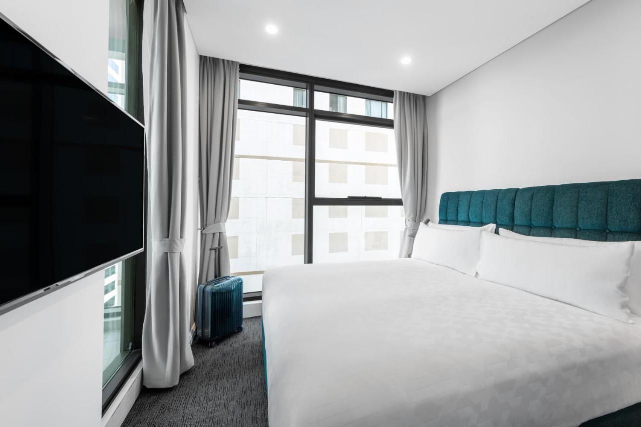 Meriton Suites Sussex Street, Сидни Екстериор снимка The photo shows a modern hotel room featuring a large bed with a light-colored duvet and a blue upholstered headboard. There are two pillows on the bed. A large window allows natural light to enter the room, and gray curtains frame the window. Next t