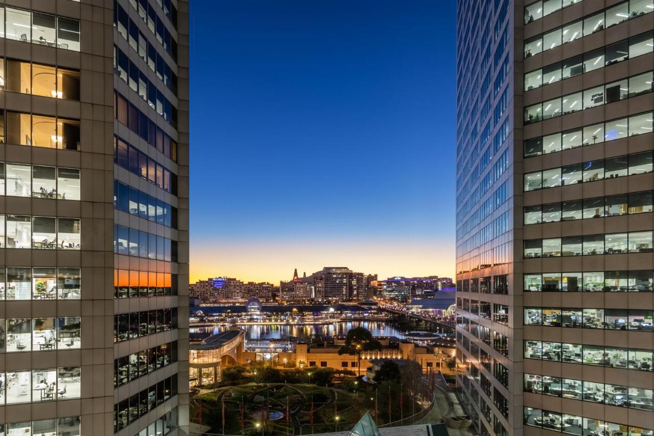 Meriton Suites Sussex Street, Сидни Екстериор снимка The photo shows a view between two tall buildings, likely office towers, during twilight. On one side, there are glass windows reflecting the evening sky that transitions from blue to orange near the horizon. In the background, a cityscape is visible