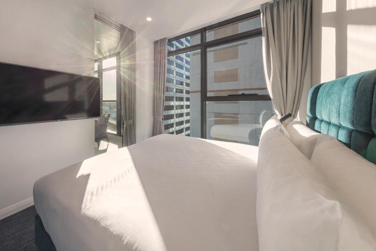 Meriton Suites Sussex Street, Сидни Екстериор снимка The photo shows a modern hotel room with a neatly made bed featuring white bedding and a plush, tufted headboard in a shade of blue. Natural light is streaming through large windows, which are covered partially by light gray curtains. A flat-screen t