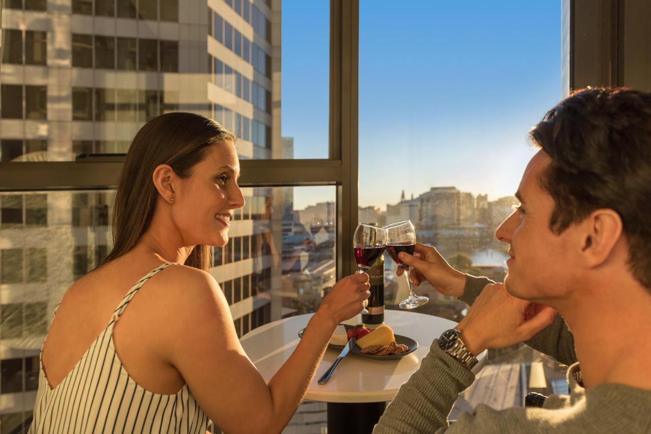 Meriton Suites Sussex Street, Сидни Екстериор снимка The photo depicts a couple enjoying a drink together in a modern setting. They are sitting at a small table near a large window, which provides a view of a cityscape and a sunset. Both individuals are smiling and engaged in a cheerful toast with glas