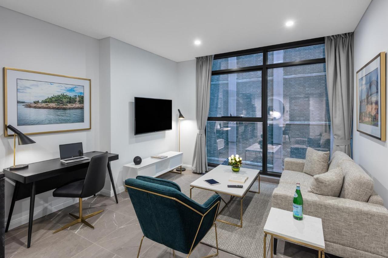 Meriton Suites Sussex Street, Сидни Екстериор снимка The photo shows a modern living area with a light color scheme. There is a comfortable seating arrangement that includes a beige sofa and an accent chair in teal. A coffee table is positioned in front of the sofa, with a small vase of flowers on it. 