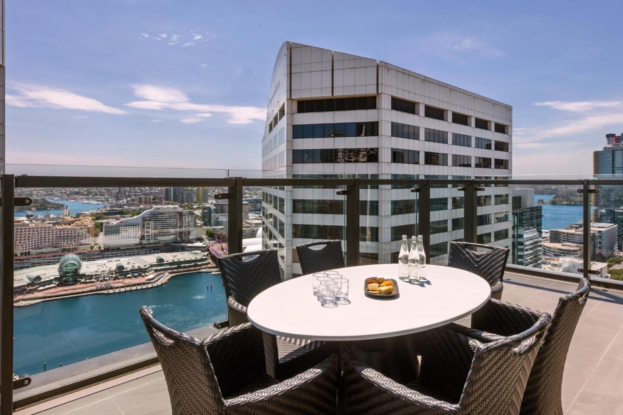 Meriton Suites Sussex Street, Сидни Екстериор снимка The photo shows a balcony or terrace with a round table and several chairs arranged around it. The table is set with some drinks and a plate, possibly containing food. In the background, there is a view of a waterway and several buildings, including 