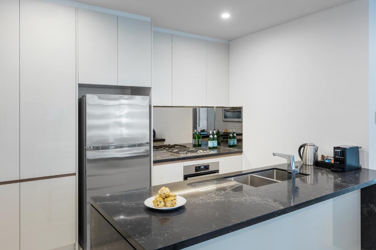 Meriton Suites Sussex Street, Сидни Екстериор снимка The photo shows a modern kitchen with a sleek, minimalist design. Key features include:

- A shiny stainless steel refrigerator on the left.
- A mirrored backsplash above the stove, which has a gas burner.
- A dark countertop made of what appears to 