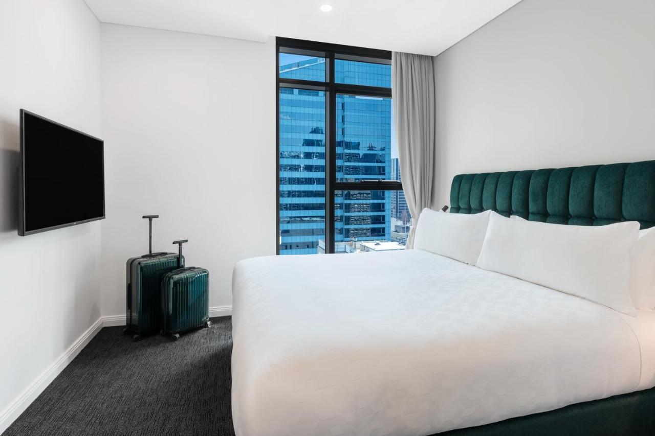 Meriton Suites Sussex Street, Сидни Екстериор снимка The photo shows a modern hotel room featuring a large bed with white bedding and a green upholstered headboard. There are two green suitcases placed beside the bed. A flat-screen TV is mounted on the wall opposite the bed. The room has a large window
