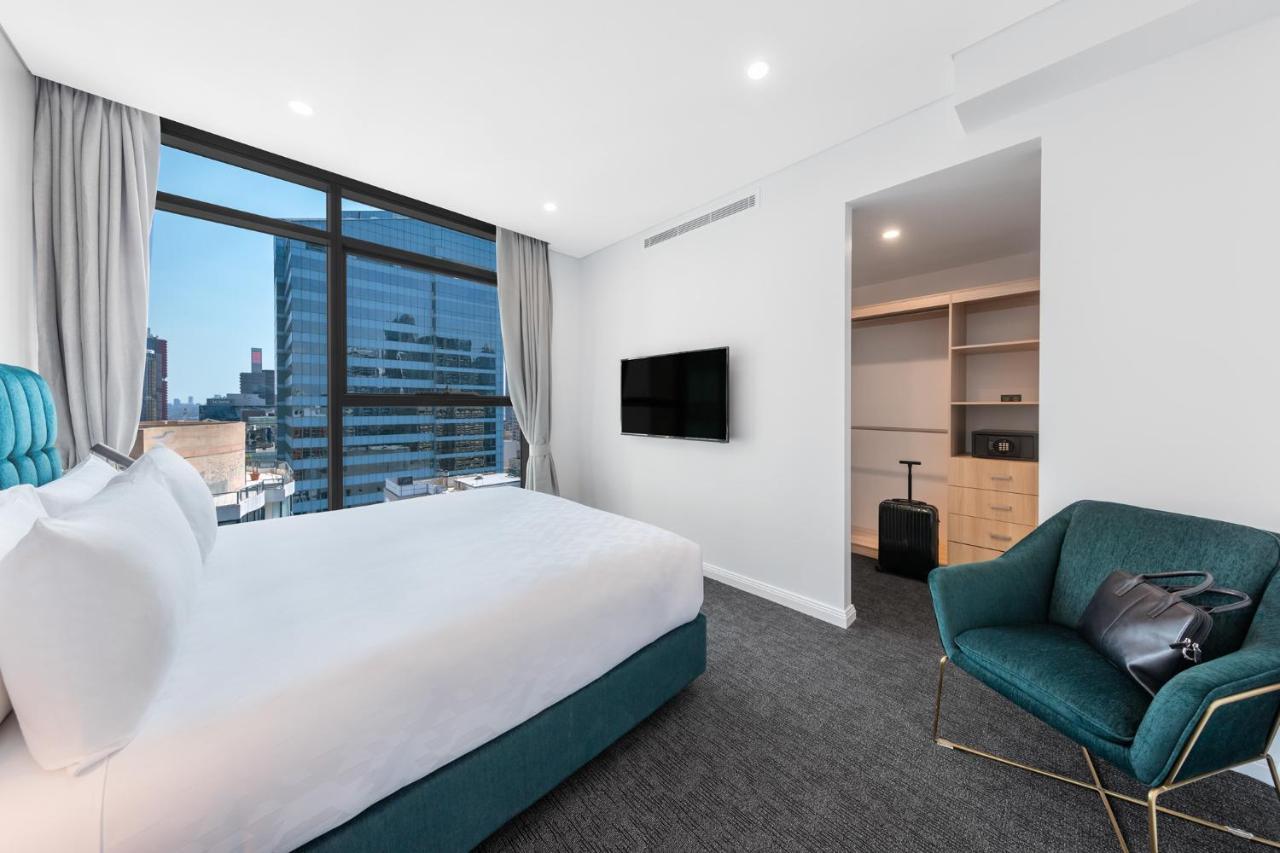 Meriton Suites Sussex Street, Сидни Екстериор снимка The photo shows a modern hotel room. It features a large window that allows natural light to fill the space, offering a view of the city skyline. The room includes a neatly made bed with a white duvet and a teal-colored base. There is a small green a