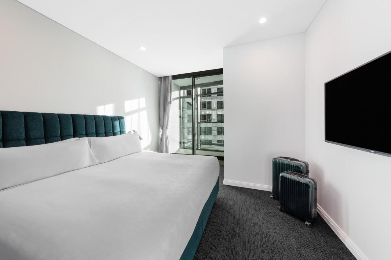 Meriton Suites Sussex Street, Сидни Екстериор снимка The photo shows a modern hotel room featuring a large bed with a headboard in a teal color. The bedding is white and appears neatly arranged. There is a large window allowing natural light to enter, offering a view of an outdoor area. A flat-screen T