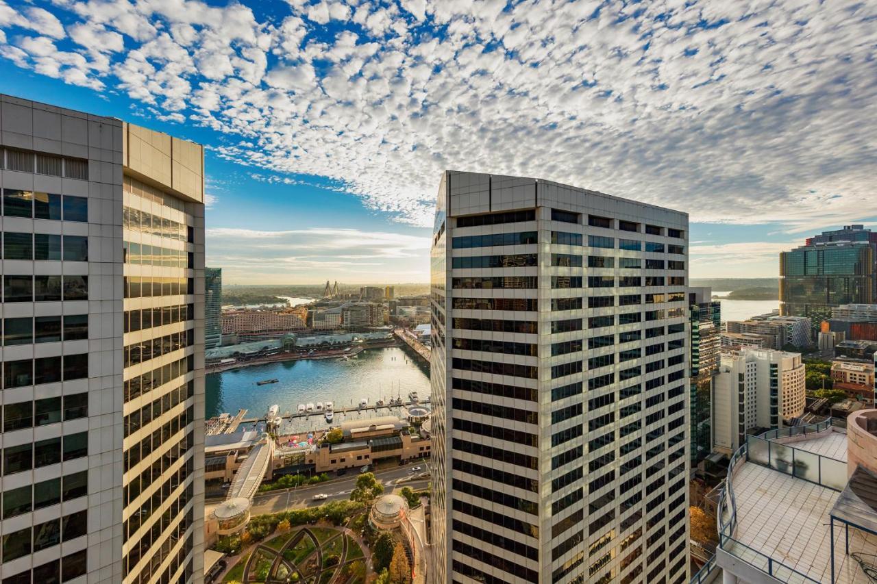 Meriton Suites Sussex Street, Сидни Екстериор снимка The photo depicts a panoramic view of a modern cityscape featuring tall commercial buildings with glass facades. The shot is taken from a height, showing the intricate architecture of the nearby structures. Below, there is a body of water, which appe