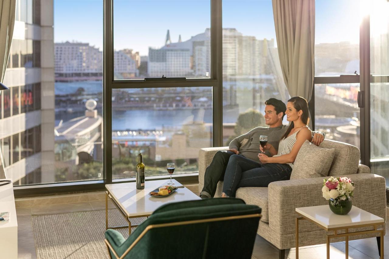 Meriton Suites Sussex Street, Сидни Екстериор снимка The photo shows a cozy indoor setting with a couple sitting closely together on a light-colored sofa. They are holding drinks, likely wine, and appear to be enjoying each other's company while looking out of a large window. The window provides a view