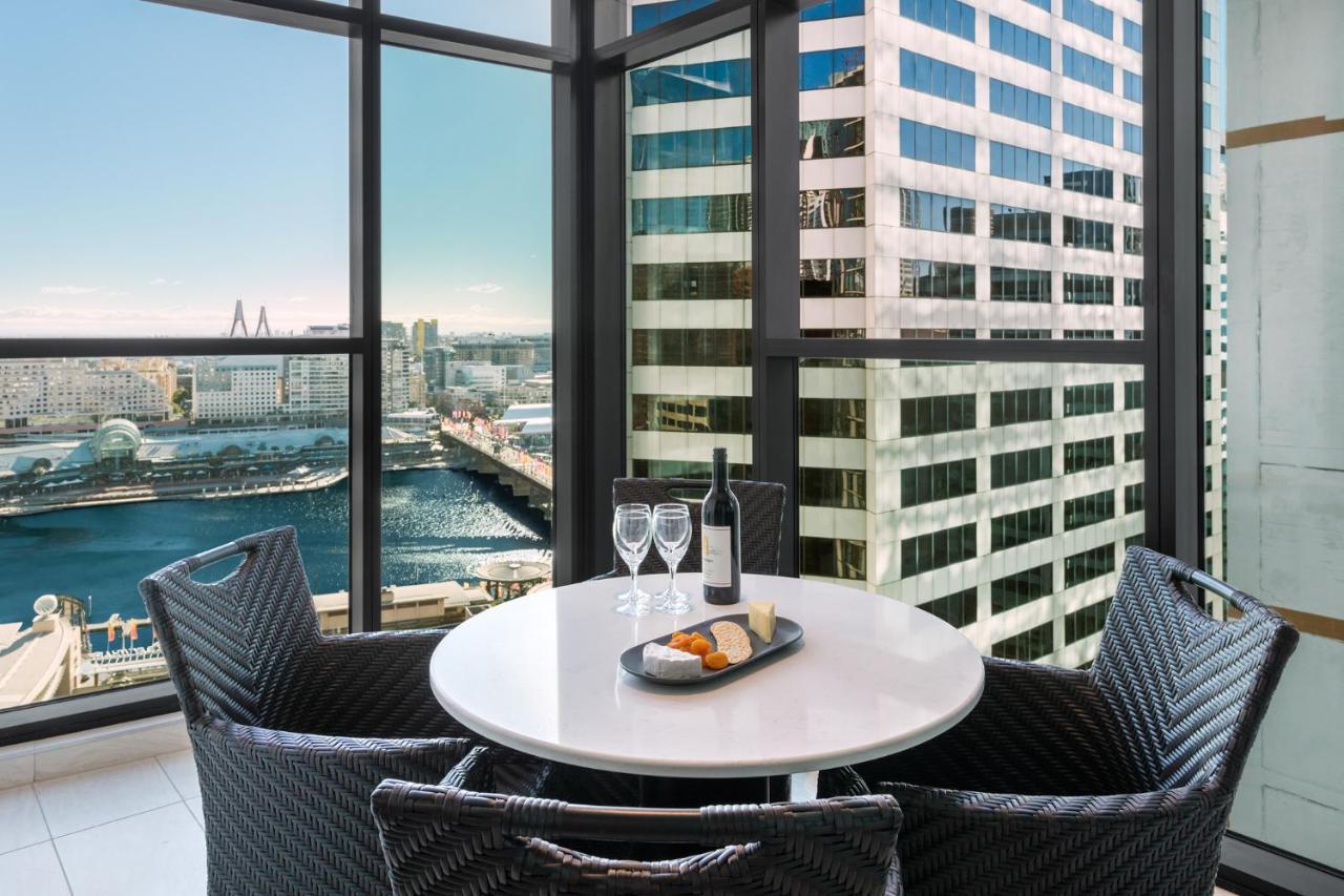 Meriton Suites Sussex Street, Сидни Екстериор снимка The photo shows a modern dining area with a round table and four woven chairs. On the table, there are two wine glasses, a bottle of wine, and a plate with food items. The large windows provide a view of a cityscape featuring tall buildings and a bod