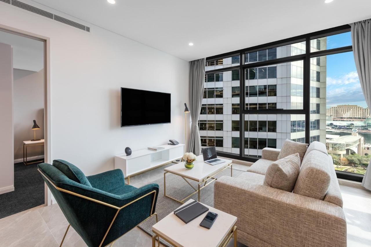 Meriton Suites Sussex Street, Сидни Екстериор снимка The photo shows a modern living room with large windows that let in natural light, providing a view of a cityscape outside. The interior features a comfortable light-colored sofa and a stylish dark green armchair. A sleek TV is mounted on the wall op