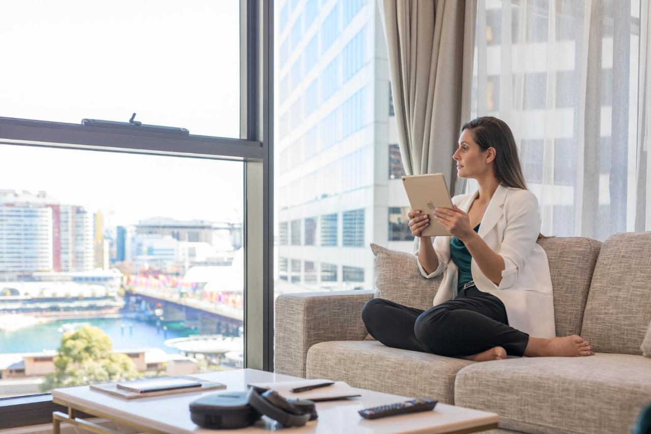 Meriton Suites Sussex Street, Сидни Екстериор снимка The image portrays a woman sitting cross-legged on a light-colored sofa, engaged with a tablet in her hands. She is dressed in a white blazer and dark pants, appearing relaxed and focused. The setting features large windows that provide a view of a w