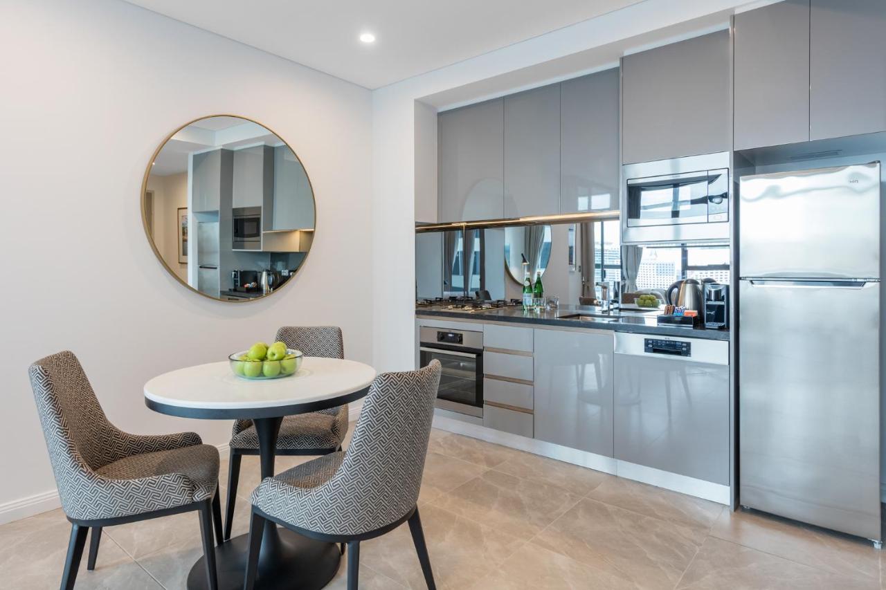 Meriton Suites Sussex Street, Сидни Екстериор снимка The photo features a modern kitchen and dining area. The kitchen has sleek grey cabinets and countertops, with built-in appliances including an oven and possibly a cooktop. There's a large stainless steel refrigerator on the right. A round dining tab