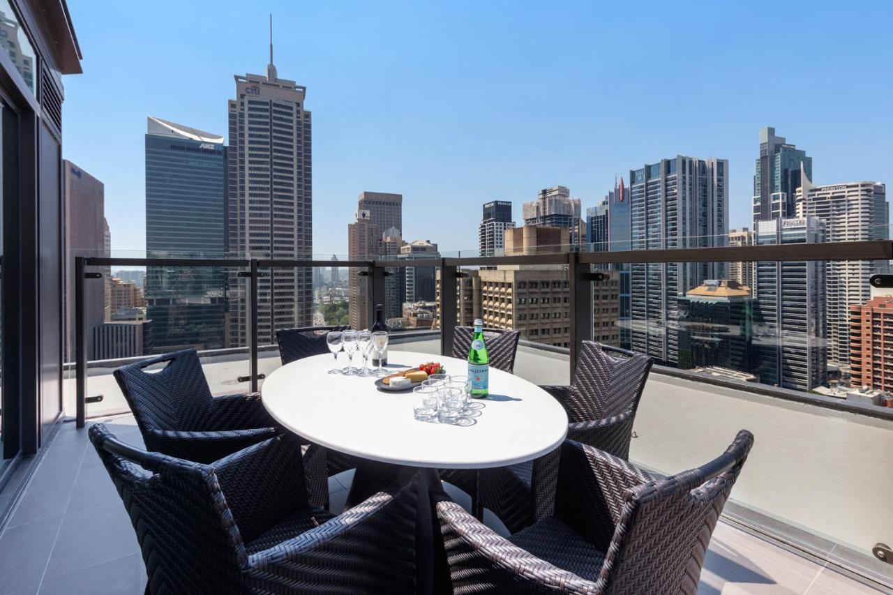 Meriton Suites Sussex Street, Сидни Екстериор снимка The photo shows a stylish outdoor dining area on a balcony with a round white table surrounded by black wicker chairs. On the table, there are several glasses, a bottle of sparkling water, and a plate of snacks. In the background, there is a panorami