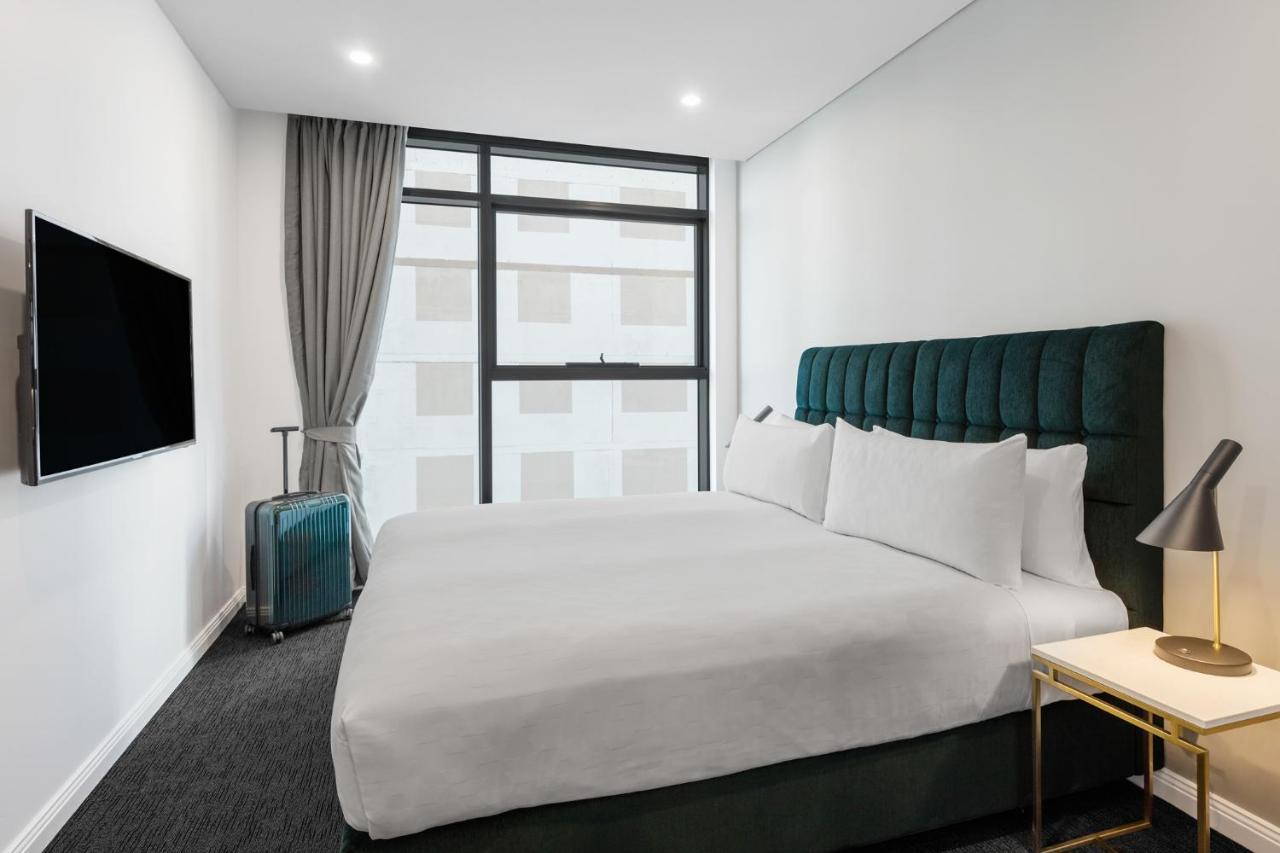 Meriton Suites Sussex Street, Сидни Екстериор снимка The photo shows a modern hotel room. It features a large bed with a dark green tufted headboard and multiple white pillows. There's a nightstand with a lamp beside the bed. A sleek television is mounted on the wall opposite the bed, and a suitcase is