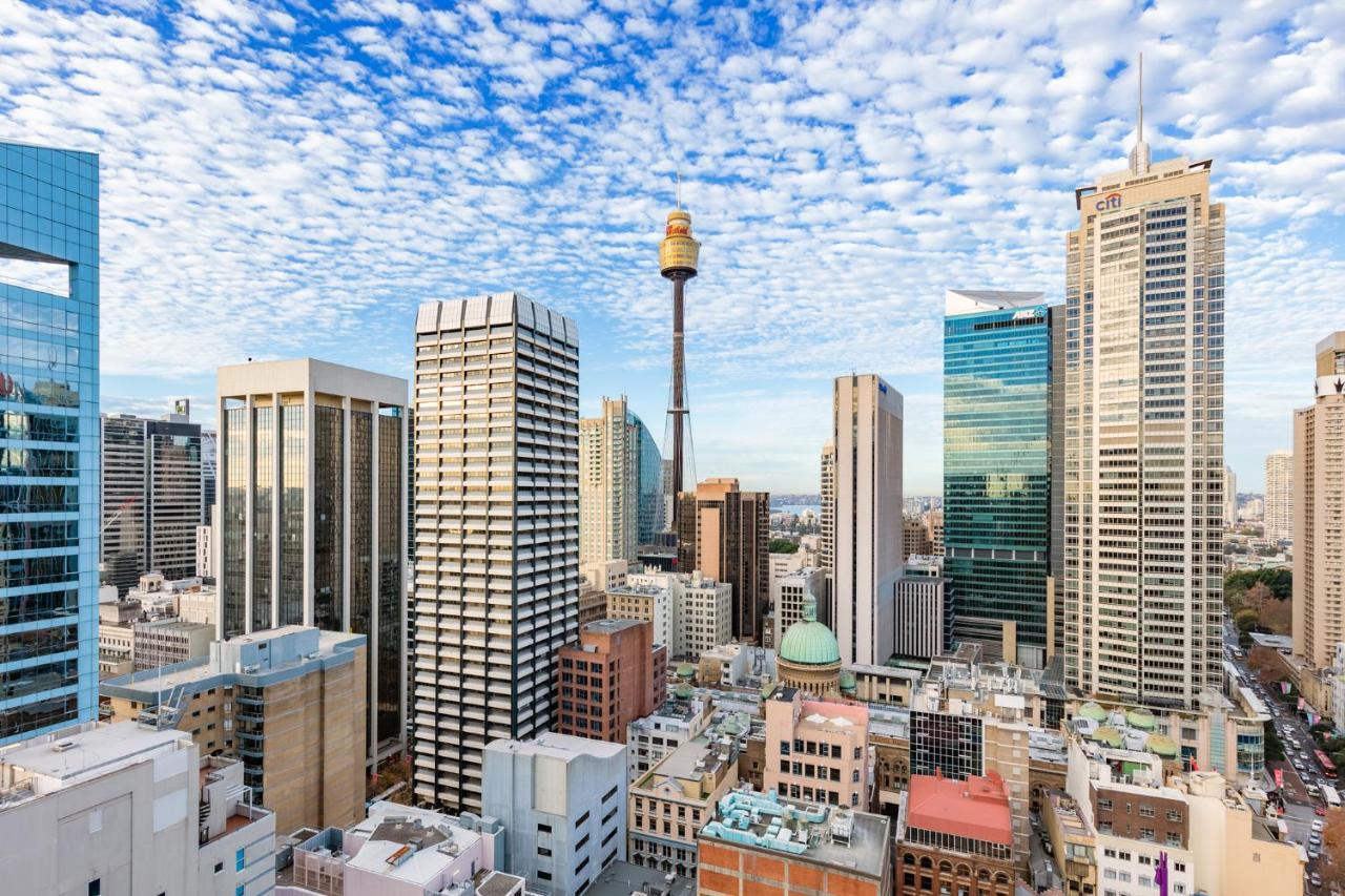 Meriton Suites Sussex Street, Сидни Екстериор снимка The photo shows a panoramic view of a city skyline dominated by tall skyscrapers. In the center, there is a notable tower with a distinctive golden structure at its top, which is likely a landmark of the city. The buildings have a mix of modern glass