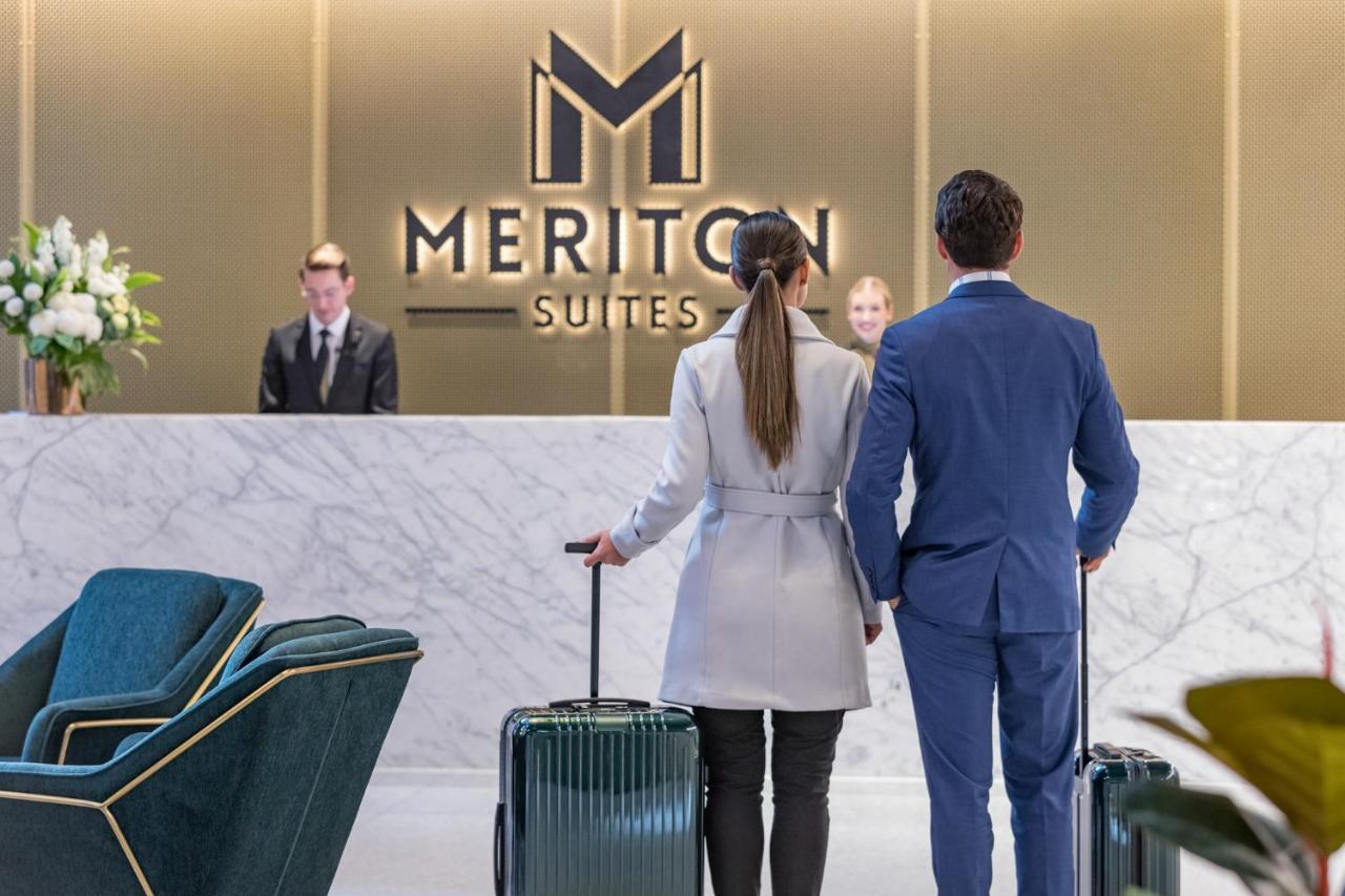 Meriton Suites Sussex Street, Сидни Екстериор снимка The photo shows a couple standing at a reception area in a hotel named "Meriton Suites." They are both facing the reception desk, where staff members are visible. The man is dressed in a suit, and the woman is wearing a light-colored coat. They each 