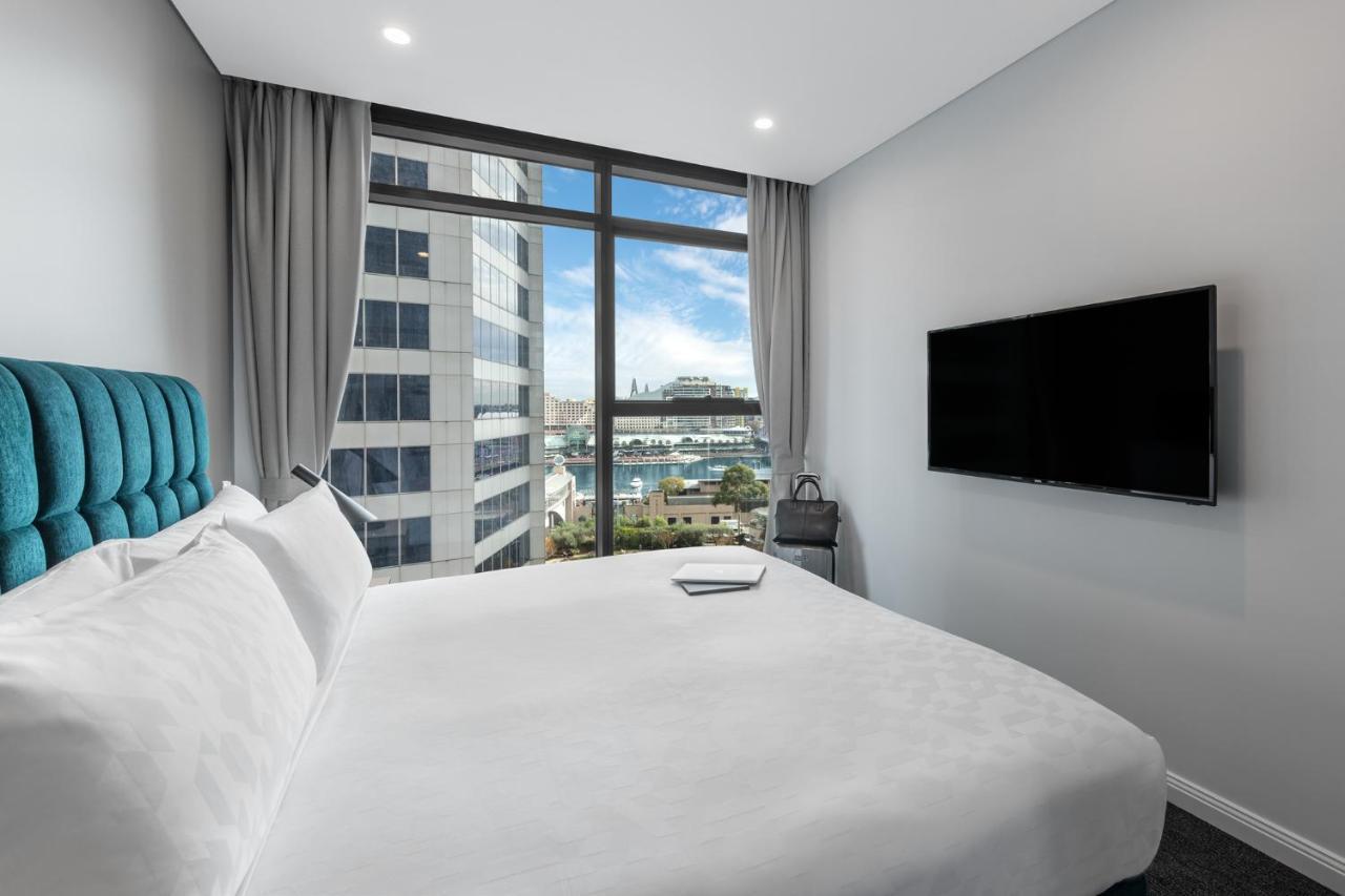 Meriton Suites Sussex Street, Сидни Екстериор снимка The photo shows a modern hotel room with a large bed featuring a white comforter and pillows. There is a blue headboard that adds a pop of color to the decor. A large window provides a view of the outside, which includes buildings and a glimpse of wa