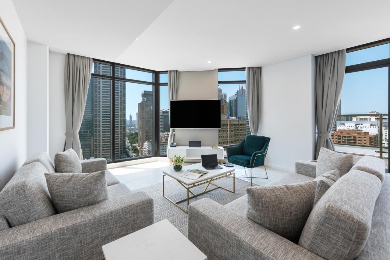 Meriton Suites Sussex Street, Сидни Екстериор снимка The photo showcases a modern living room with large windows offering a view of a city skyline. The room features a light color palette with gray sofas arranged around a coffee table. A flat-screen TV is mounted on the wall, and there is a green armch