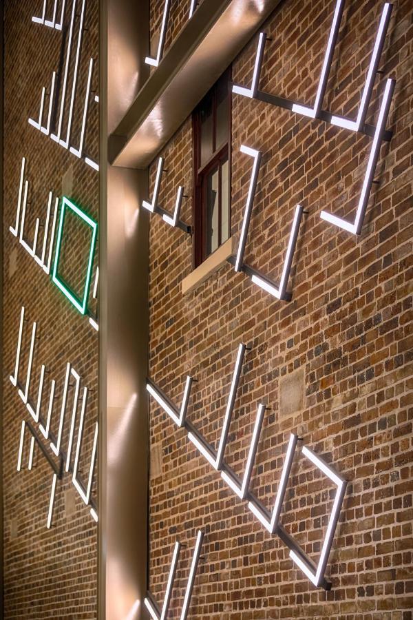 Meriton Suites Sussex Street, Сидни Екстериор снимка The photo shows a brick wall featuring a design made up of linear light fixtures. These light strips create geometric shapes and patterns, with some sections illuminated in white and one section highlighted in green. The combination of the brick text