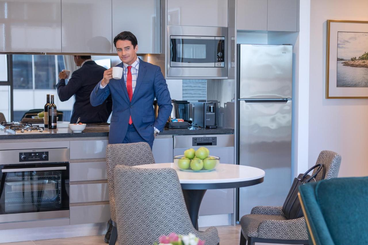Meriton Suites Sussex Street, Сидни Екстериор снимка The photo depicts a well-dressed man in a suit standing in a modern kitchen. He is holding a cup, possibly of coffee, and appears to be looking towards the camera. The kitchen features contemporary appliances, including a microwave and refrigerator. 
