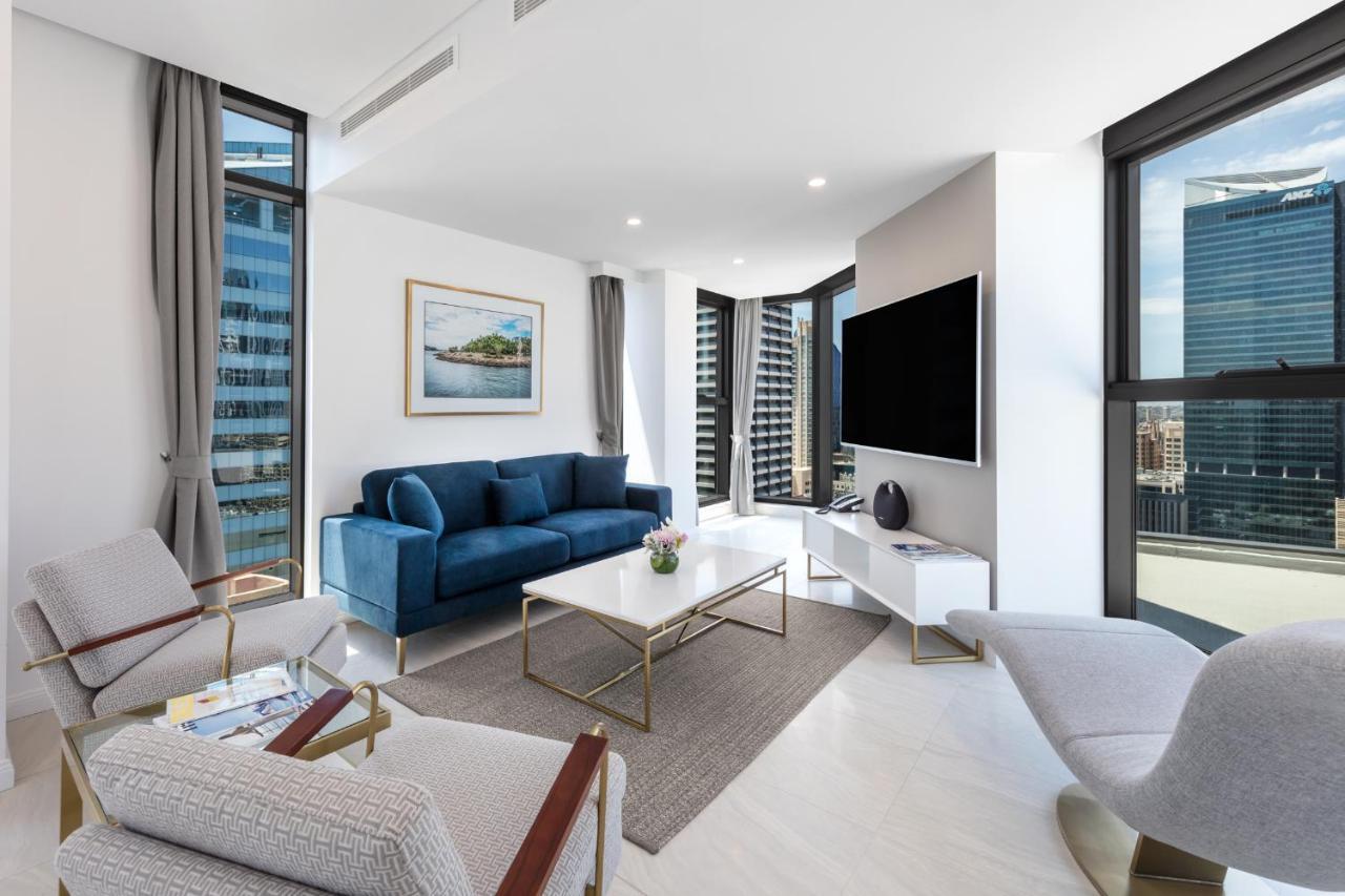 Meriton Suites Sussex Street, Сидни Екстериор снимка The photo shows a modern living room featuring large windows that allow plenty of natural light. The room is elegantly designed with a blue sofa, a light-colored coffee table, and two armchairs. There’s a flat-screen TV mounted on the wall, and a pie